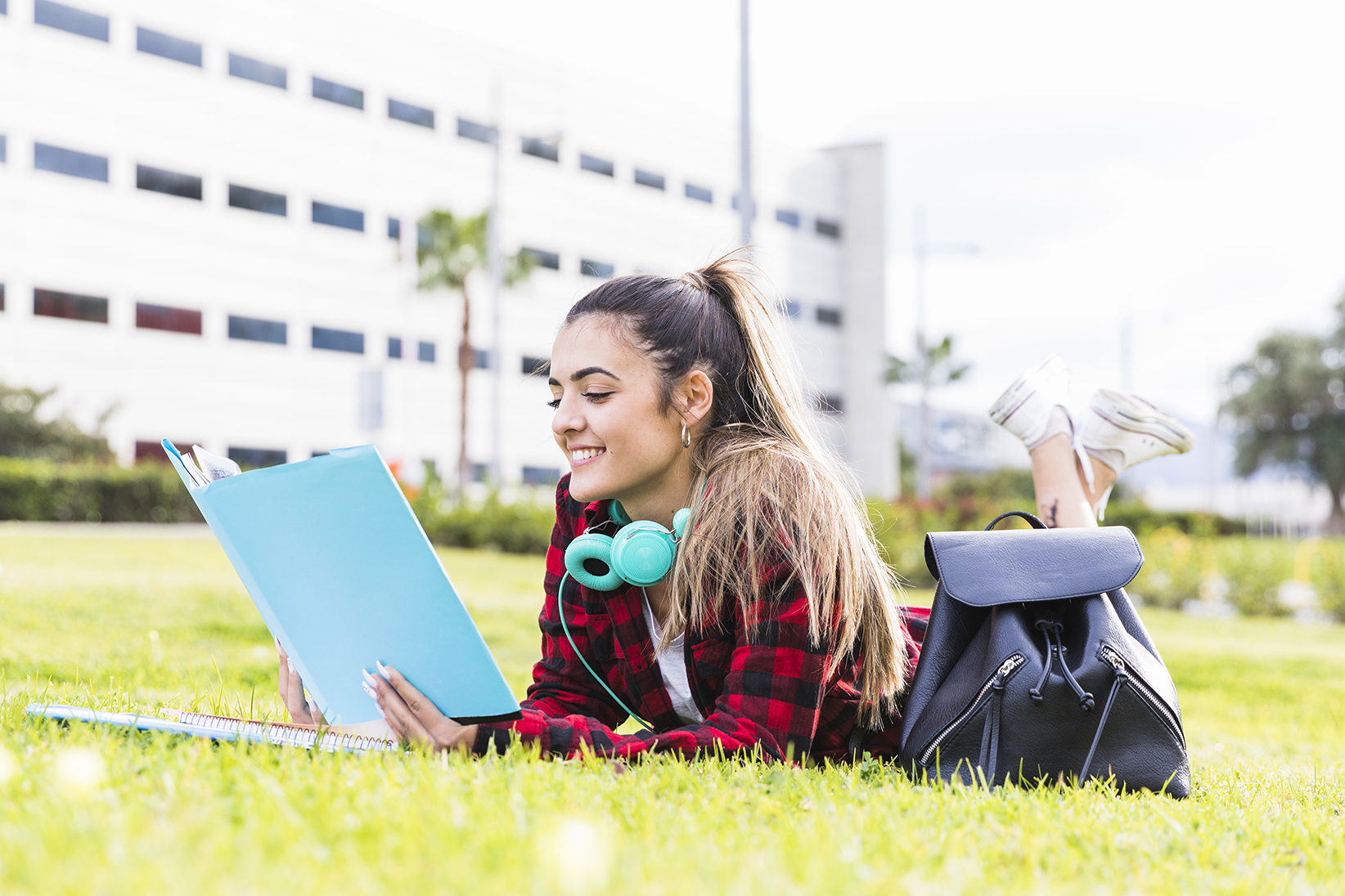 Uma jovem mulher sorrindo deitada no gramado, lendo o livro no campus da Universidade